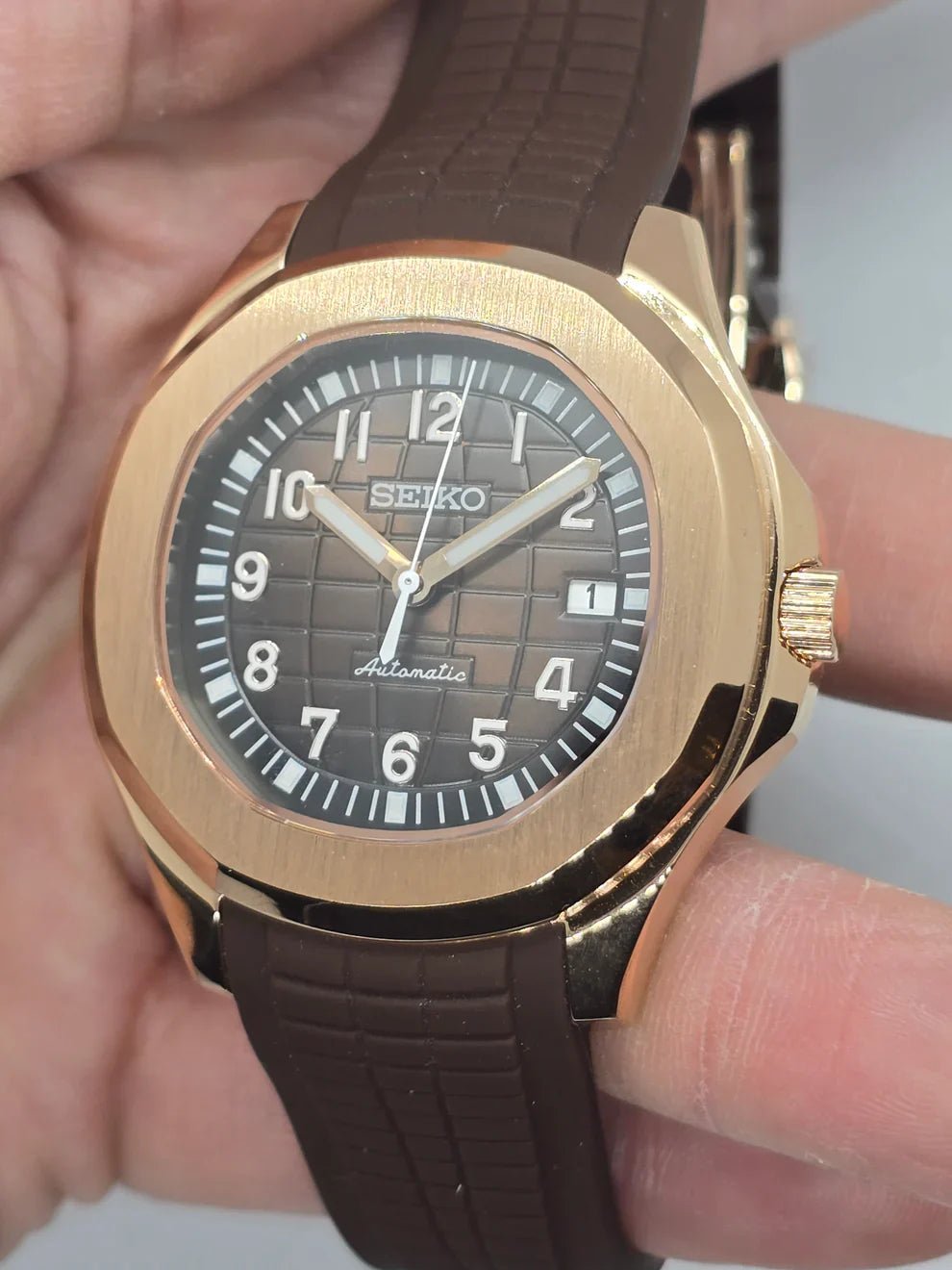 Montre automatique de style Aquanaut — configuration aux tons or rose, cadran marron (NH35) avec couronne striée aux tons or rose, en biais dans une main