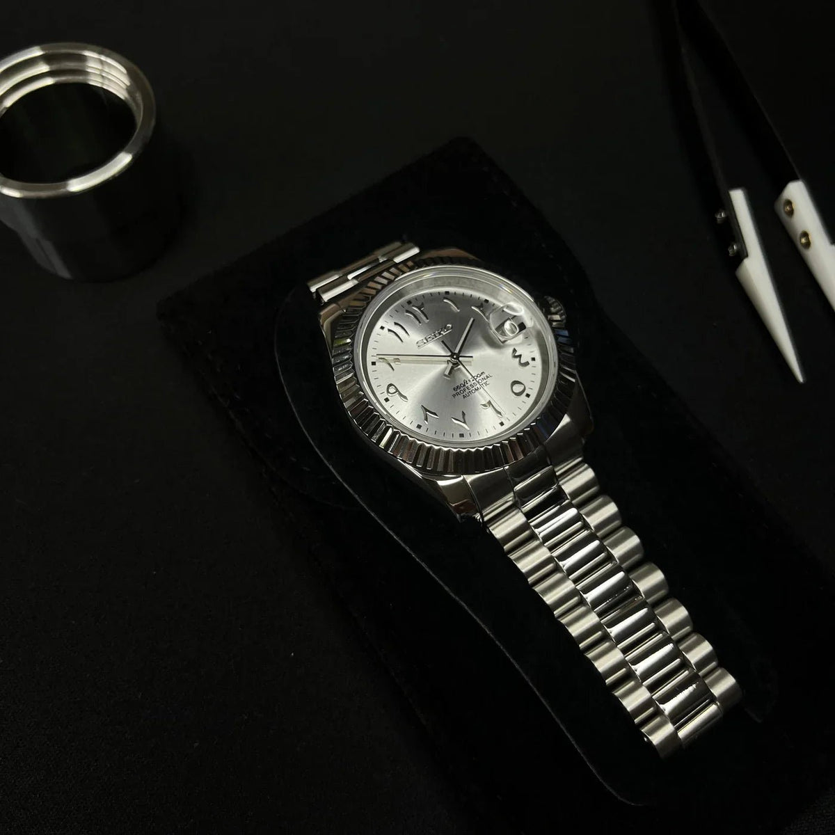 Montre automatique de style Datejust — configuration cadran argenté à chiffres arabes (NH35) avec maillons arrondis aux tons argentés, à côté d'une pince sur une pochette