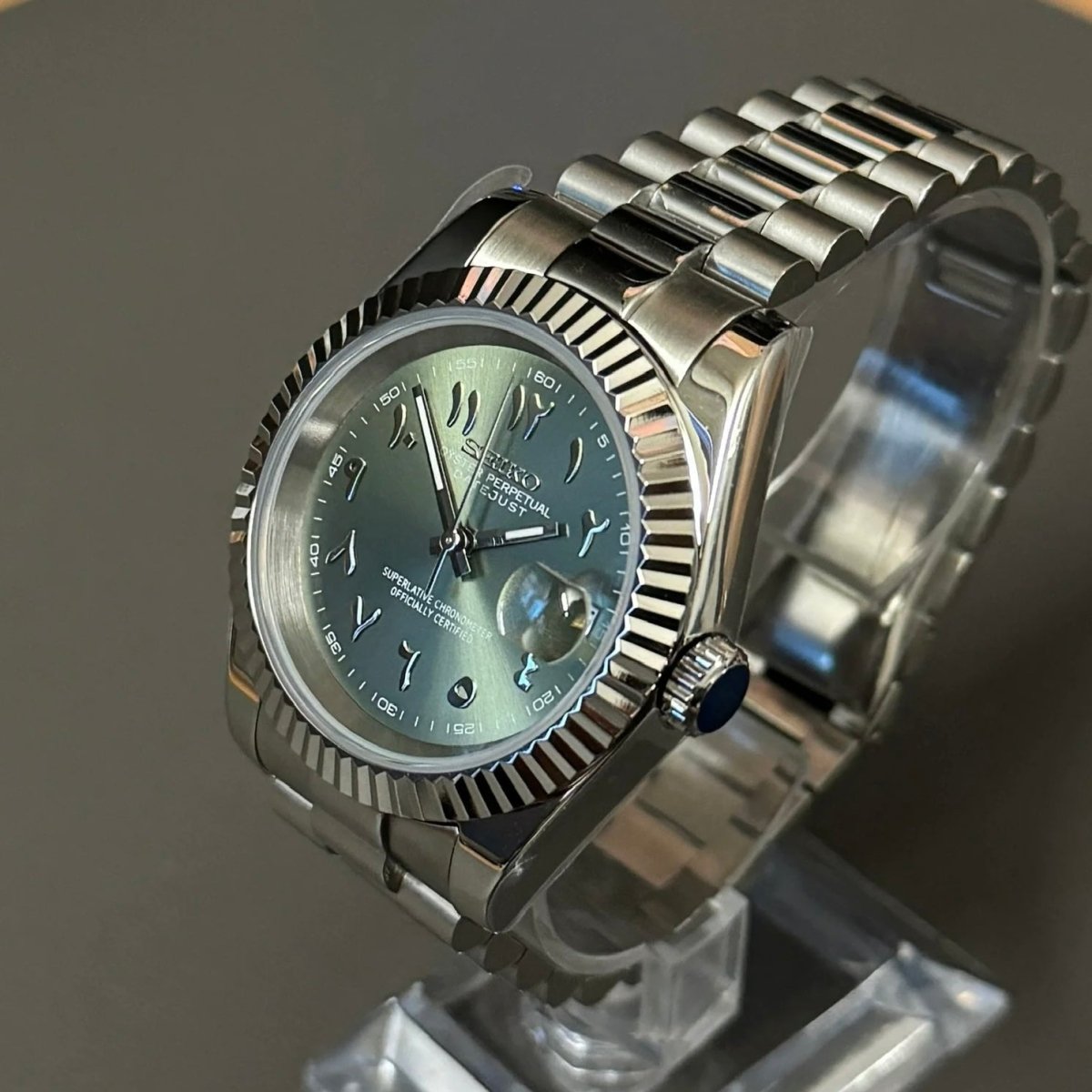 Montre automatique de style Datejust — configuration cadran vert olive à chiffres arabes (NH35) avec couronne striée aux tons argentés, vue en biais côté droit sur un support
