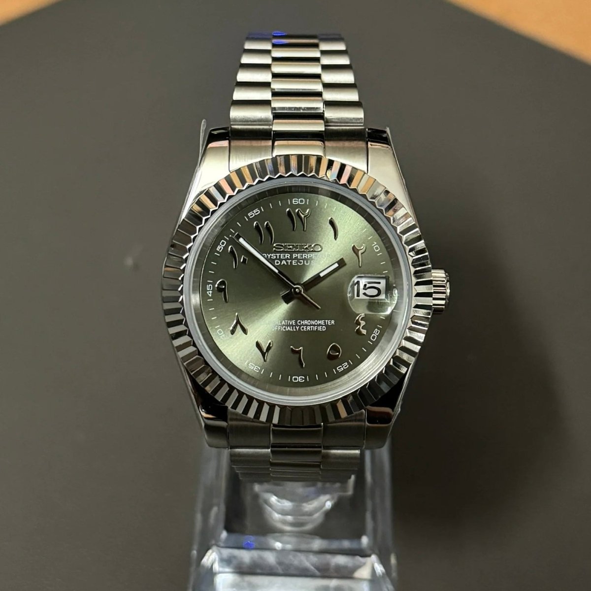 Montre automatique de style Datejust — configuration cadran vert olive à chiffres arabes (NH35) avec lunette cannelée aux tons argentés, vue de face sur un support