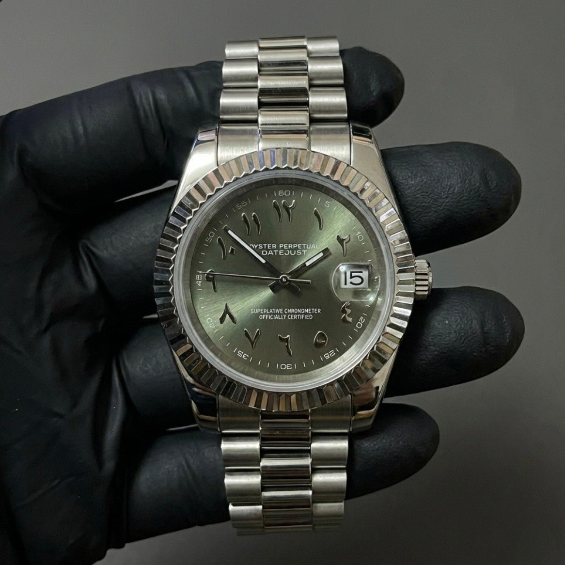 Montre automatique de style Datejust — configuration cadran vert olive à chiffres arabes (NH35) avec lunette cannelée aux tons argentés, tenue dans un gant noir