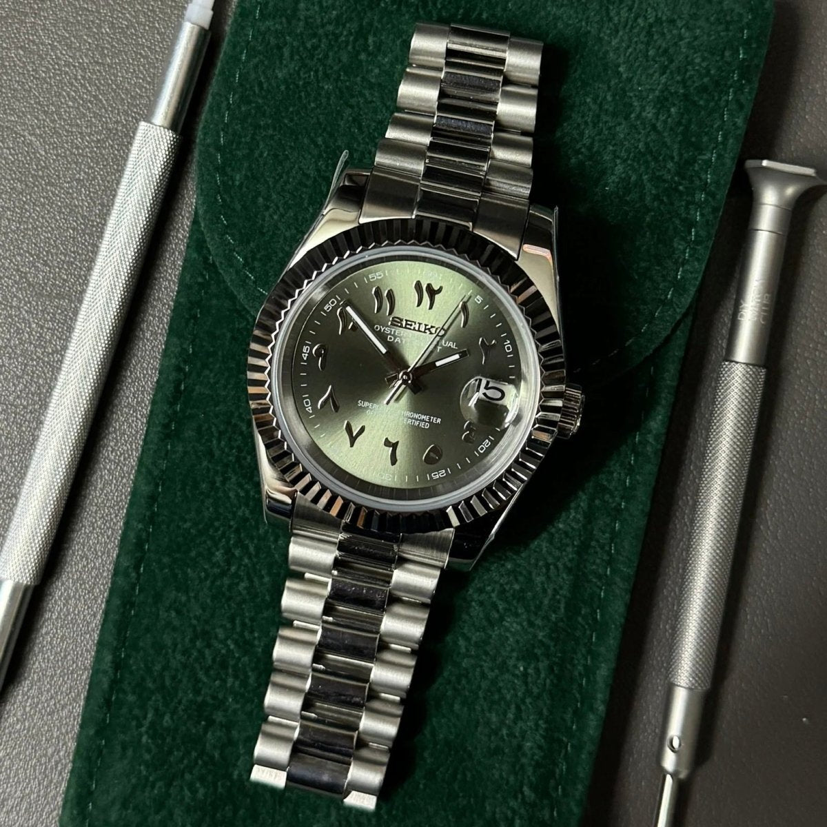 Montre automatique de style Datejust — configuration cadran vert olive à chiffres arabes (NH35) avec maillons arrondis aux tons argentés, sur une pochette verte près d'outils