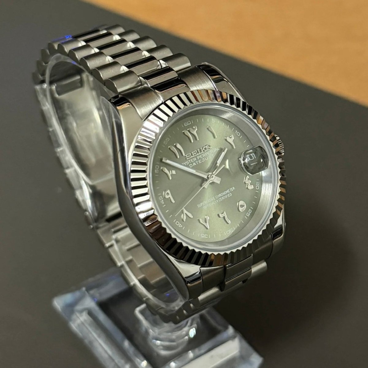 Montre automatique de style Datejust — configuration cadran vert olive à chiffres arabes (NH35) avec maillons centraux polis aux tons argentés, vue en biais côté gauche sur un support