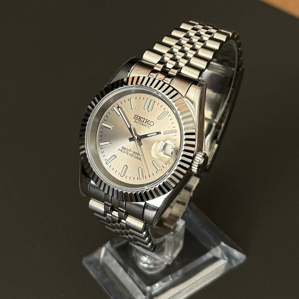 Montre automatique de style Datejust — configuration argentée, cadran champagne (NH35) avec couronne aux tons argentés, vue en biais côté droit sur un support