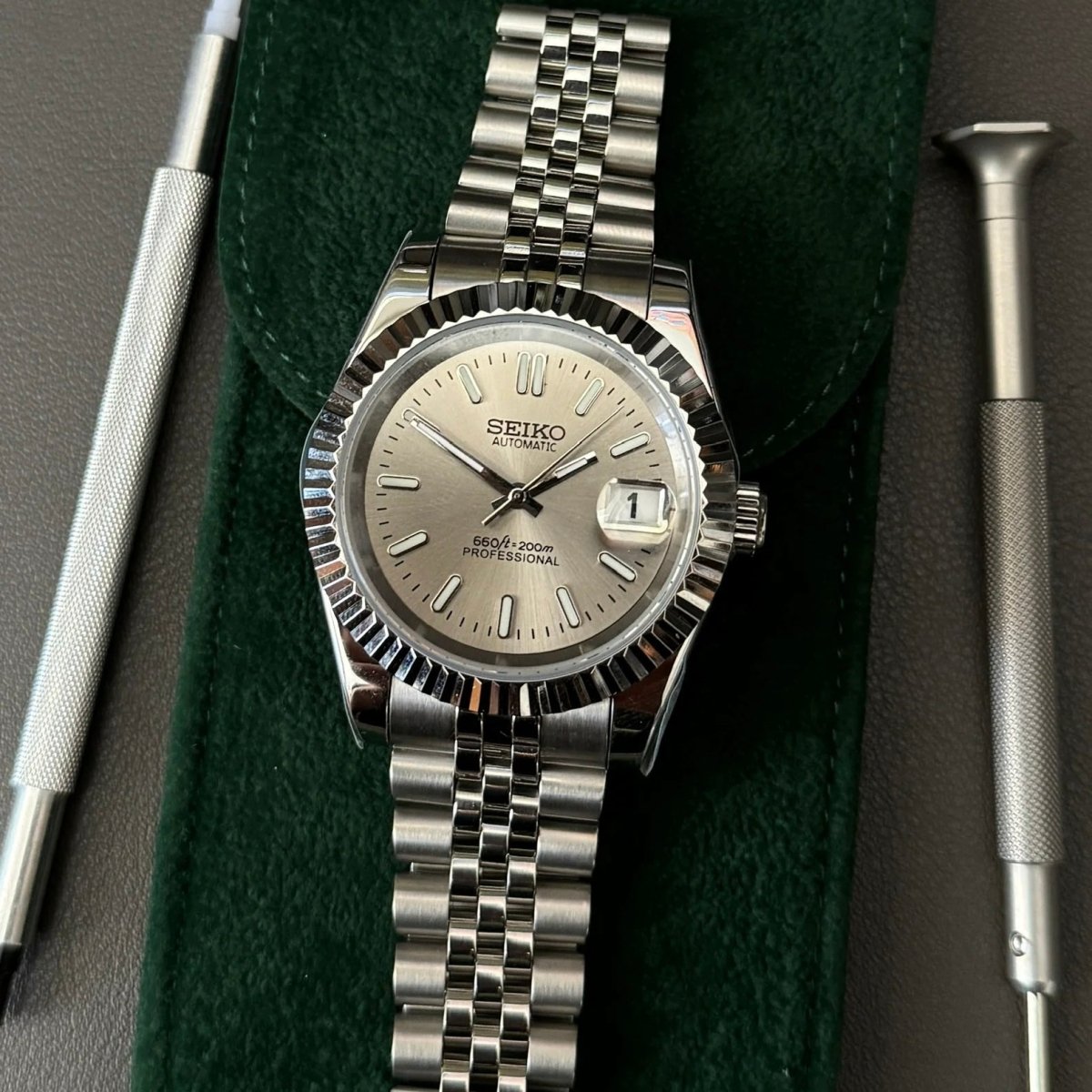 Montre automatique de style Datejust — configuration argentée, cadran champagne (NH35) avec maillons sur plusieurs rangées aux tons argentés, à côté d'outils sur une pochette verte