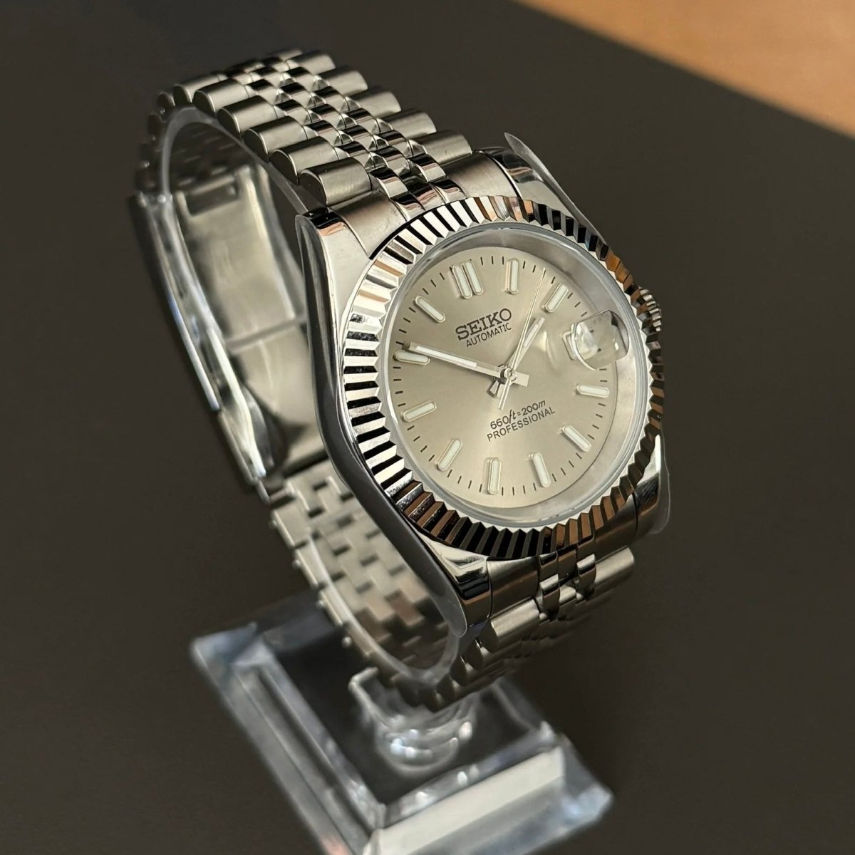 Montre automatique de style Datejust — configuration argentée, cadran champagne (NH35) avec stries de la lunette aux tons argentés, vue en biais côté gauche sur un support