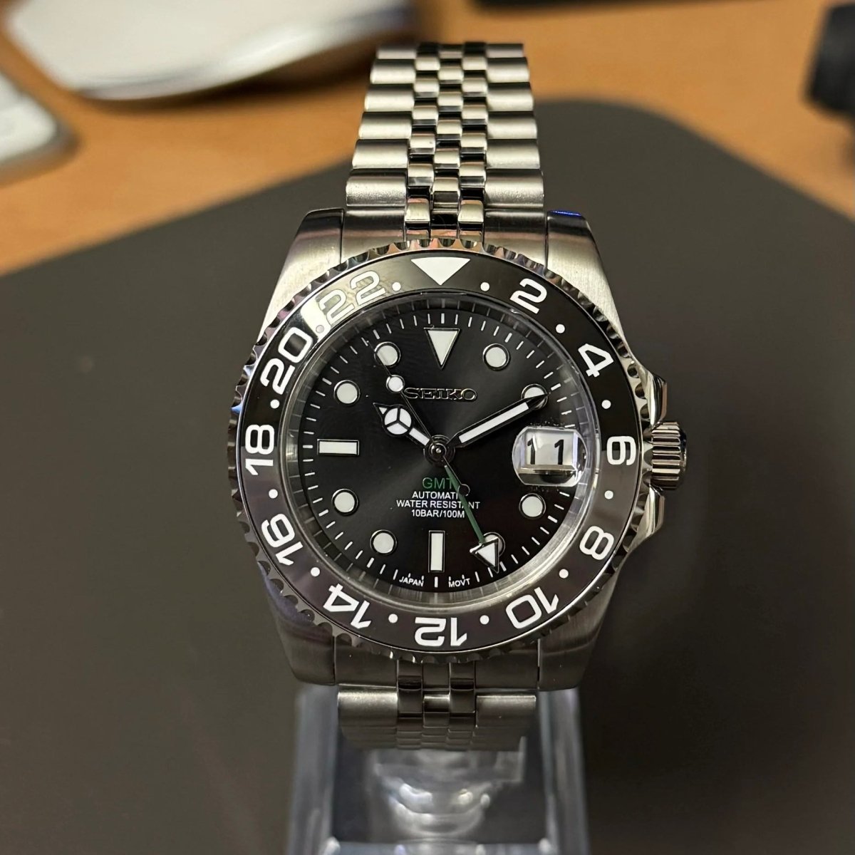 Montre automatique GMT — configuration «Bruce Wayne» (NH34) avec cadran noir et aiguille GMT verte, vue de face sur un support transparent