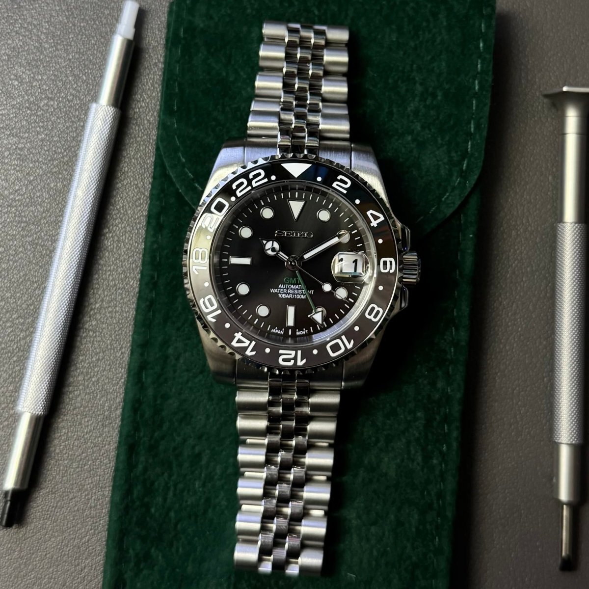 Montre automatique GMT — configuration «Bruce Wayne» (NH34) avec un cadran noir et un bracelet de style Jubilee sur une pochette verte à côté d'outils