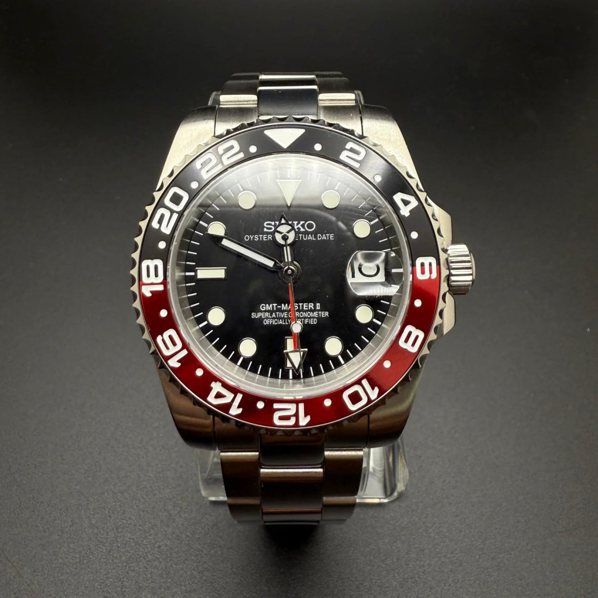 Montre automatique GMT — configuration «Coke» (NH34) avec cadran noir et date agrandie, vue de face sur un support transparent