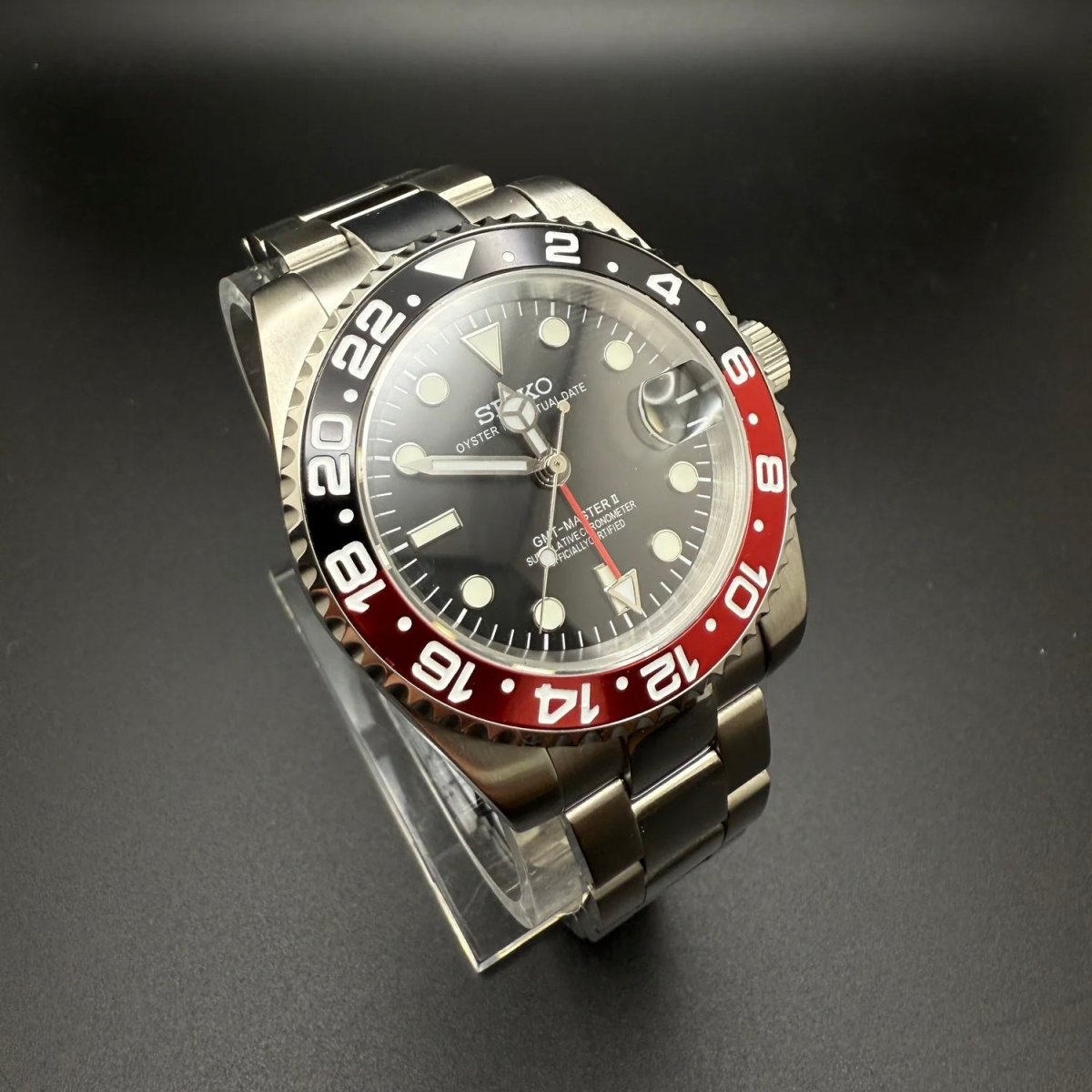 Montre automatique GMT — configuration «Coke» (NH34) avec bracelet aux tons argentés et aiguille GMT rouge, vue de trois quarts côté gauche