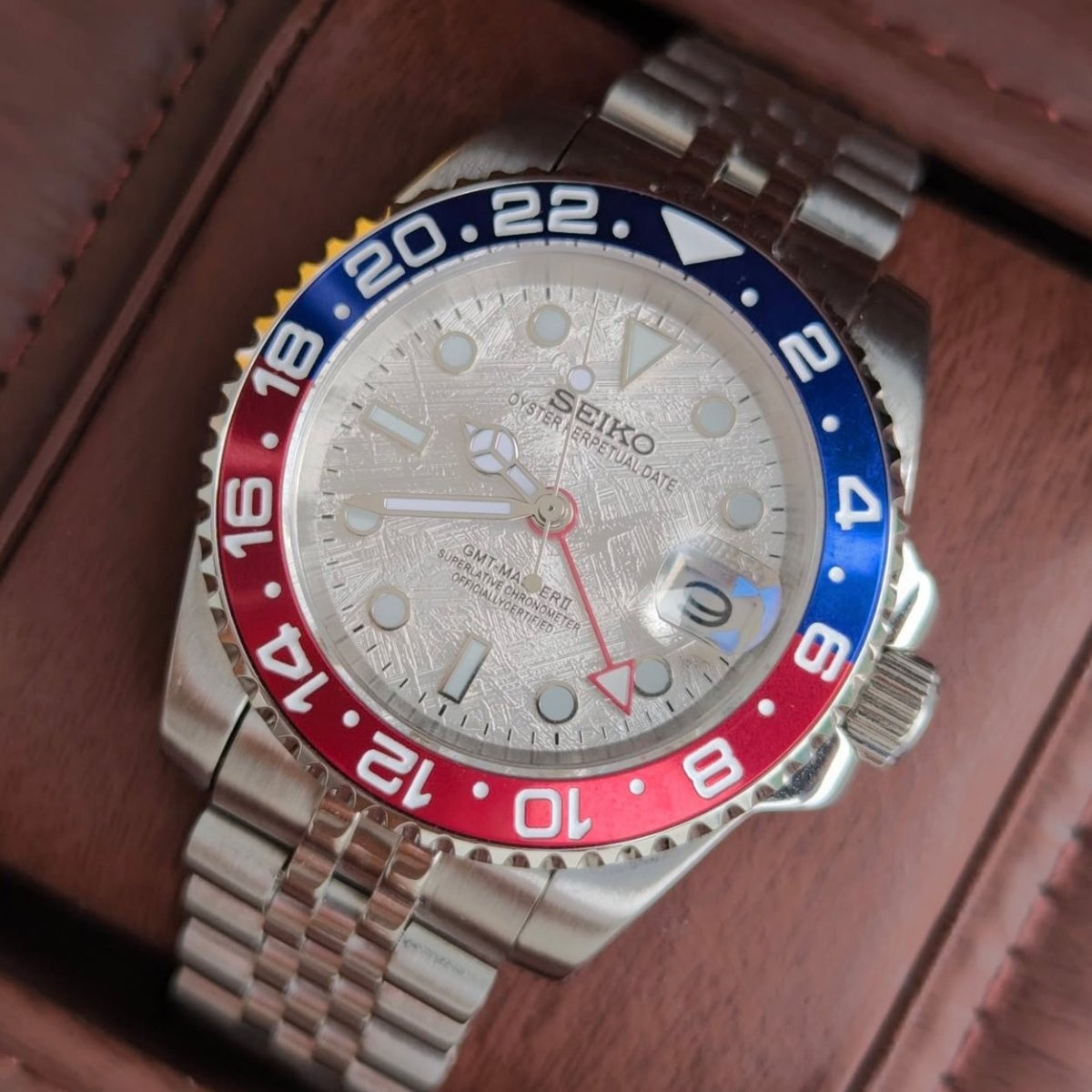 Montre automatique GMT — configuration «Meteorite» (NH34) avec cadran argenté et loupe de date, gros plan en biais dans une boîte marron
