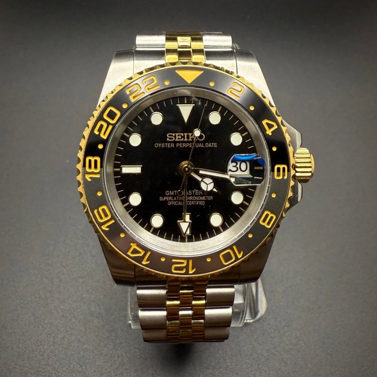 Montre automatique GMT — configuration bicolore et noire (NH34) avec lunette noire et chiffres aux tons dorés, vue de face sur un support