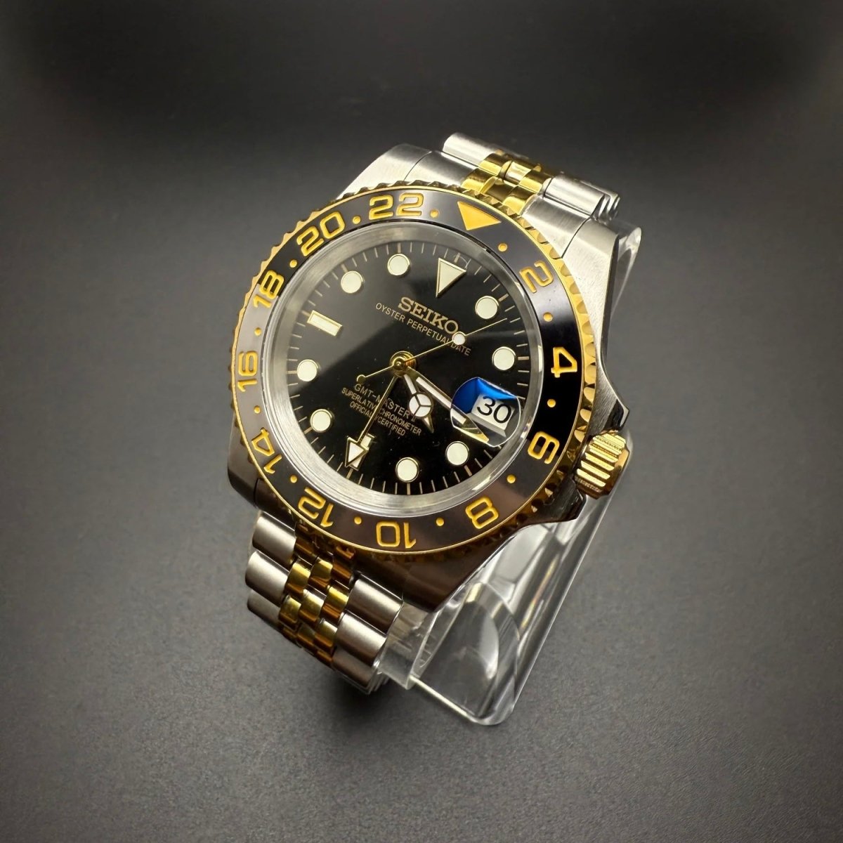 Montre automatique GMT — configuration bicolore et noire (NH34) avec cadran noir et couronne aux tons dorés, vue en biais sur un support transparent