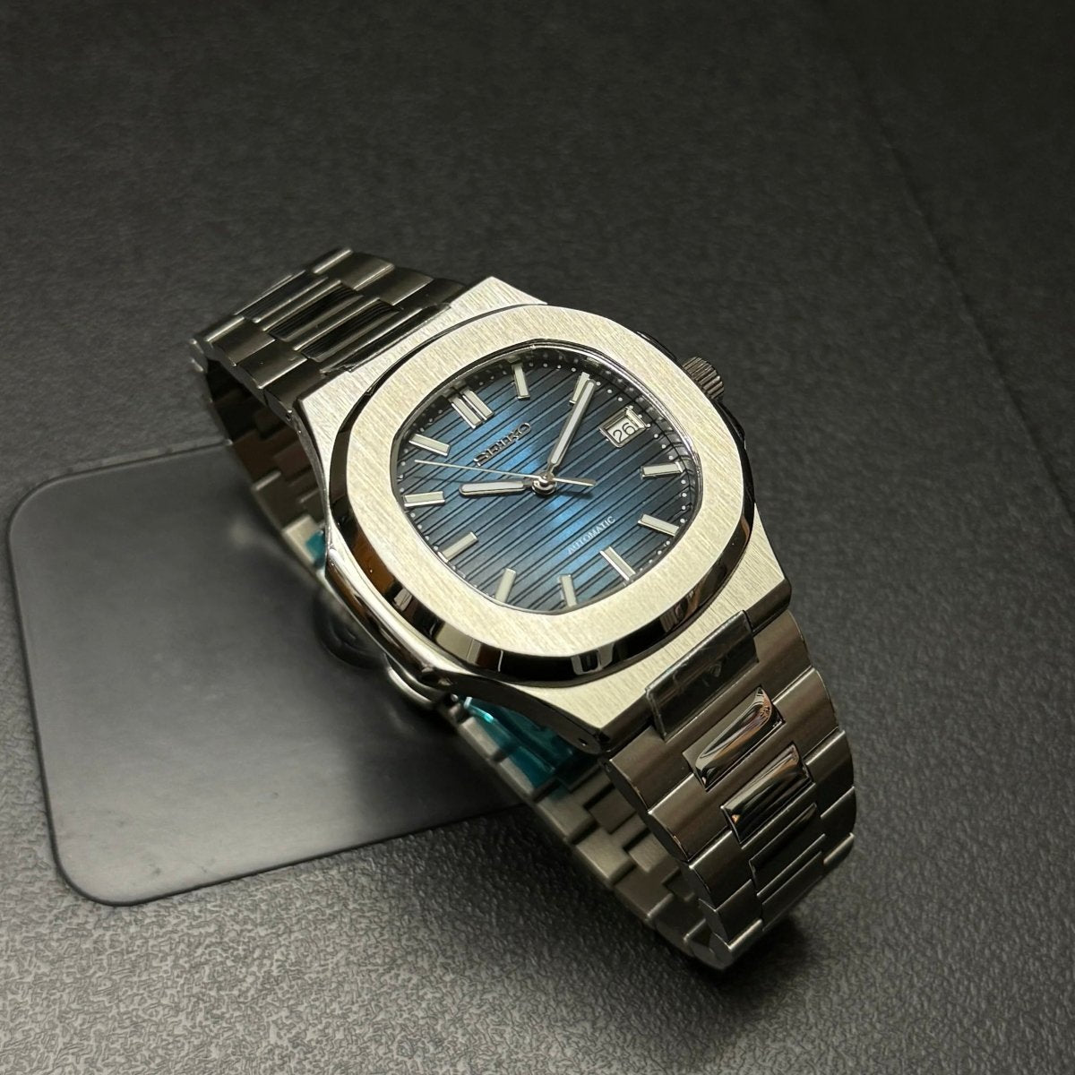 Montre automatique de style Nautilus — configuration argentée, cadran bleu (NH35) avec maillons aux tons argentés, guichet de date, sur un coussinet sombre